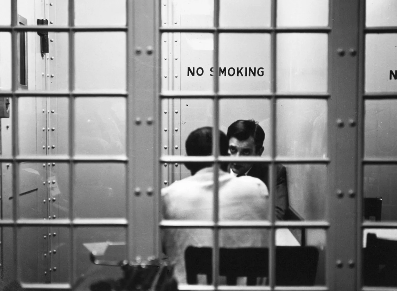 Seen through the bars and glass of a jail visiting room marked No Smoking, a lawyer leans in to talk with a client