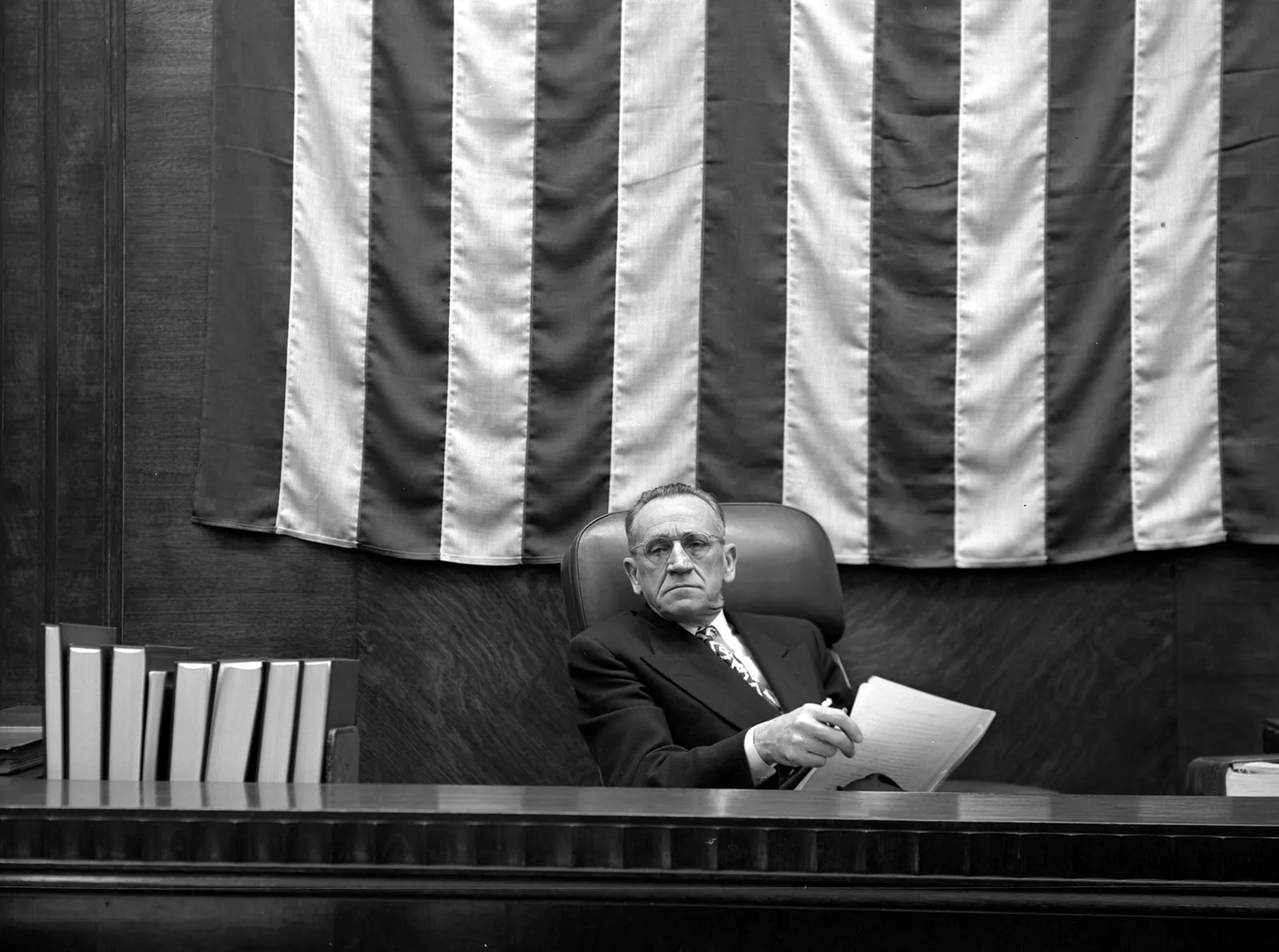 A judge sits at the bench beneath a huge draped American flag, holding papers and looking out at the courtroom